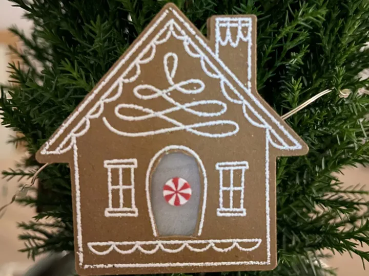 Ein aus braunem Papier geschnitztes Haus mit weißer Zuckerguss-Optik, dekoriert mit einem roten und weißen Streifen-Muster an der Tür. Das Stück hängt als Ornament an einer grünen Weihnachtsbaumzweig.