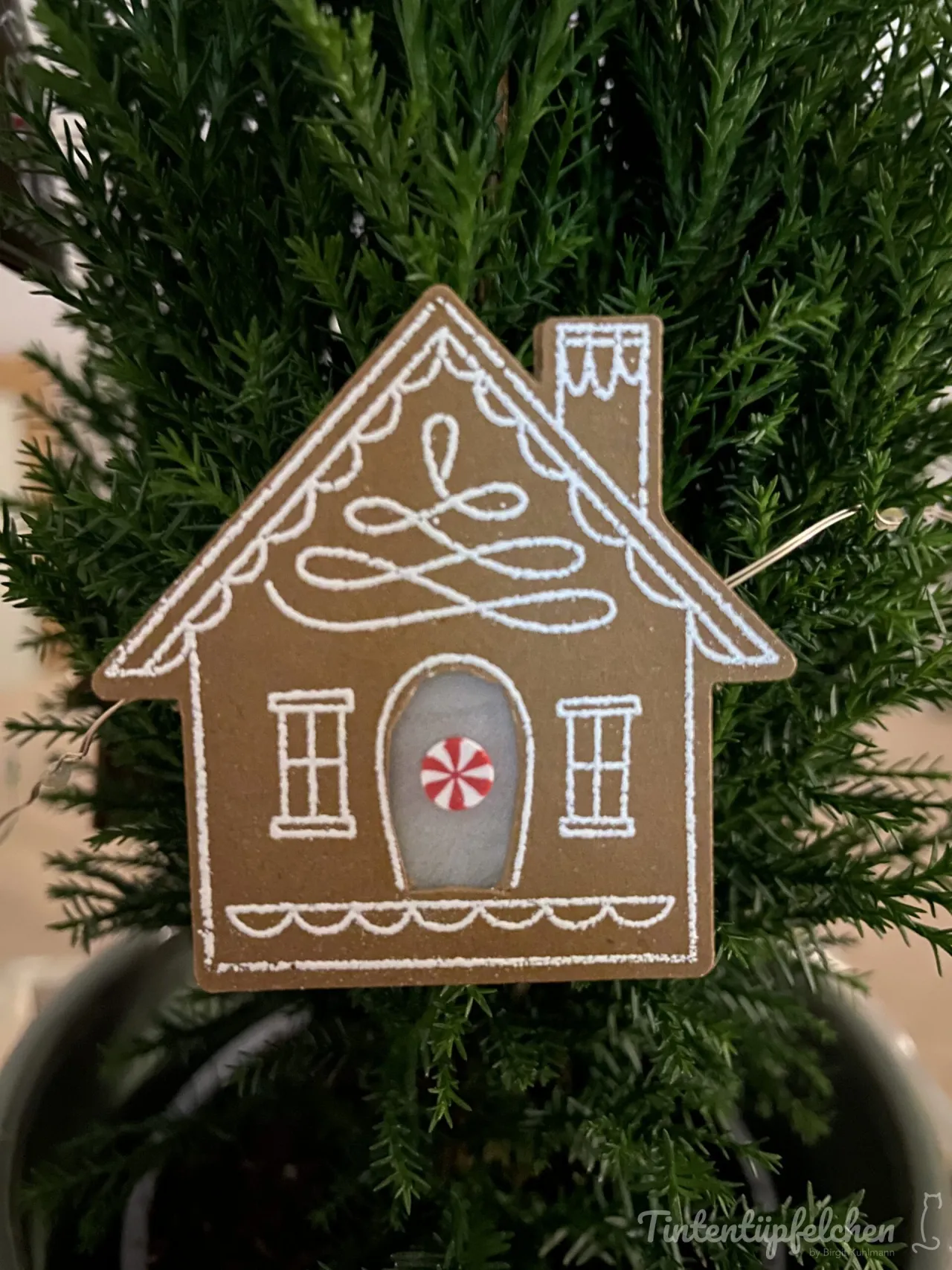 Ein aus braunem Papier geschnitztes Haus mit weißer Zuckerguss-Optik, dekoriert mit einem roten und weißen Streifen-Muster an der Tür. Das Stück hängt als Ornament an einer grünen Weihnachtsbaumzweig.
