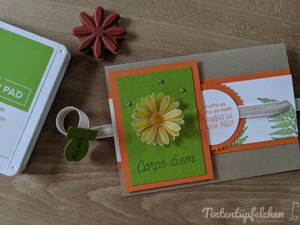 Eine selbstgebastelte Karte mit gestempeltem gelbem Gänseblümchen auf grünem Papier, umrandet von orangefarbenem Karton und einem weißen Kreis mit Text. Daneben liegen ein grüner Inkpad, ein roter Stempel in Form einer Blume und eine kleine Beschriftung aus Papier.