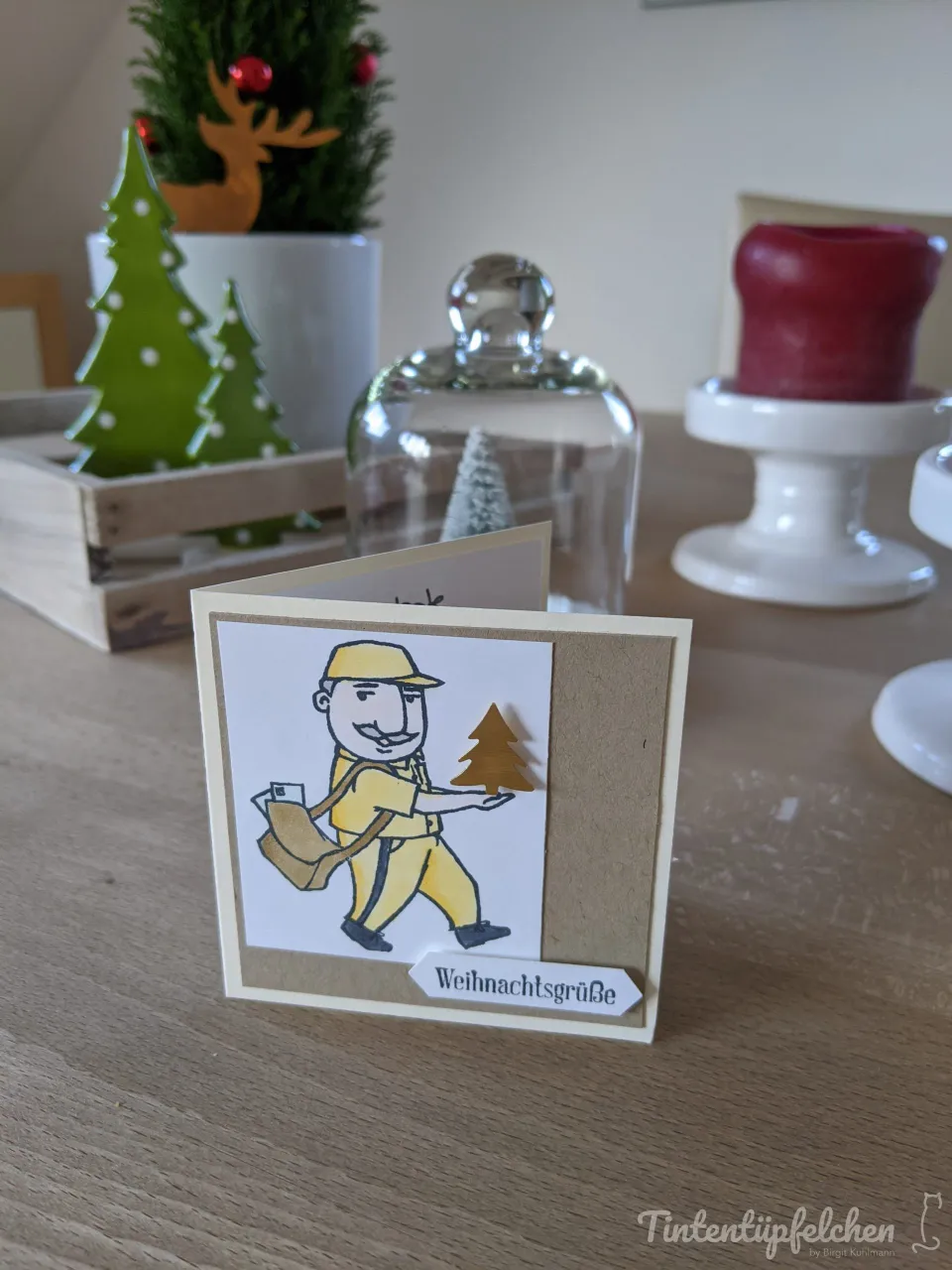 Eine Klappkarte mit einem gezeichneten Postboten in gelber Uniform, der einen kleinen goldenen Weihnachtsbaum hält. Darunter steht "Weihnachtsgrüße" auf einem weißen Fähnchen.