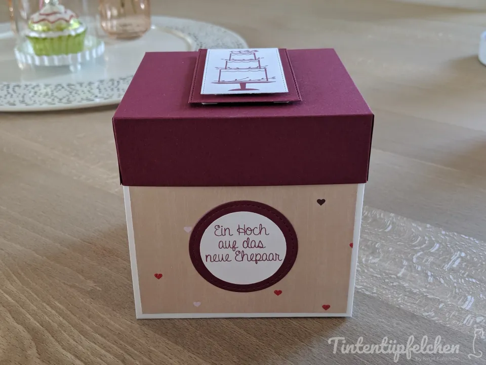Eine kastenförmige Geschenkverpackung in Bordeauxrot und Beige mit Herzen und einem Kreis mit der Aufschrift "Ein Hoch auf das neue Ehepaar". Auf dem Deckel ist ein gestempelter Hochzeitstorten-Aufleger.