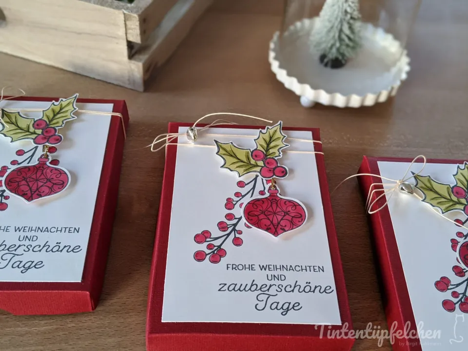 Drei rote Geschenkboxen sind mit weißen Papierbögen verziert. Auf jedem Bogen ist ein Weihnachtsmotiv mit Mistelzweigen, roten Beeren und einem roten Christbaumkugel-Anhänger gestempelt. Darunter steht der Gruß "Frohe Weihnachten und zauberschöne Tage".