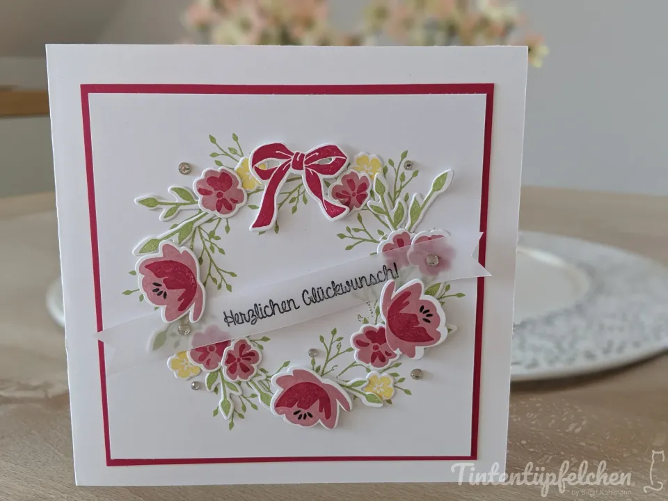Eine weiße Klappkarte mit einem Kranz aus rosa und gelben Blüten sowie grünen Blättern. Ein rotes Schleifenband und ein Banner mit der Aufschrift "Herzlichen Glückwunsch!" sind ebenfalls Teil des Kranzes.