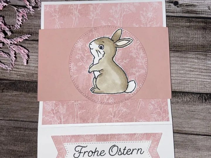 Eine Osterkarte mit gestempeltem Hasenbild in beige und rosa auf Holzoptik, umgeben von getrocknetem Zweig. Der Text „Frohe Ostern“ ist in schwarzer Schrift auf einem weißen Band mit rosa Rand platziert.