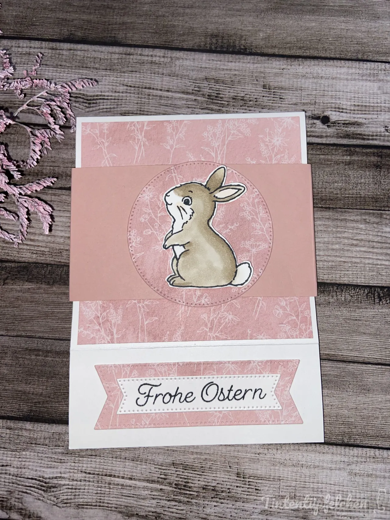 Eine Osterkarte mit gestempeltem Hasenbild in beige und rosa auf Holzoptik, umgeben von getrocknetem Zweig. Der Text „Frohe Ostern“ ist in schwarzer Schrift auf einem weißen Band mit rosa Rand platziert.