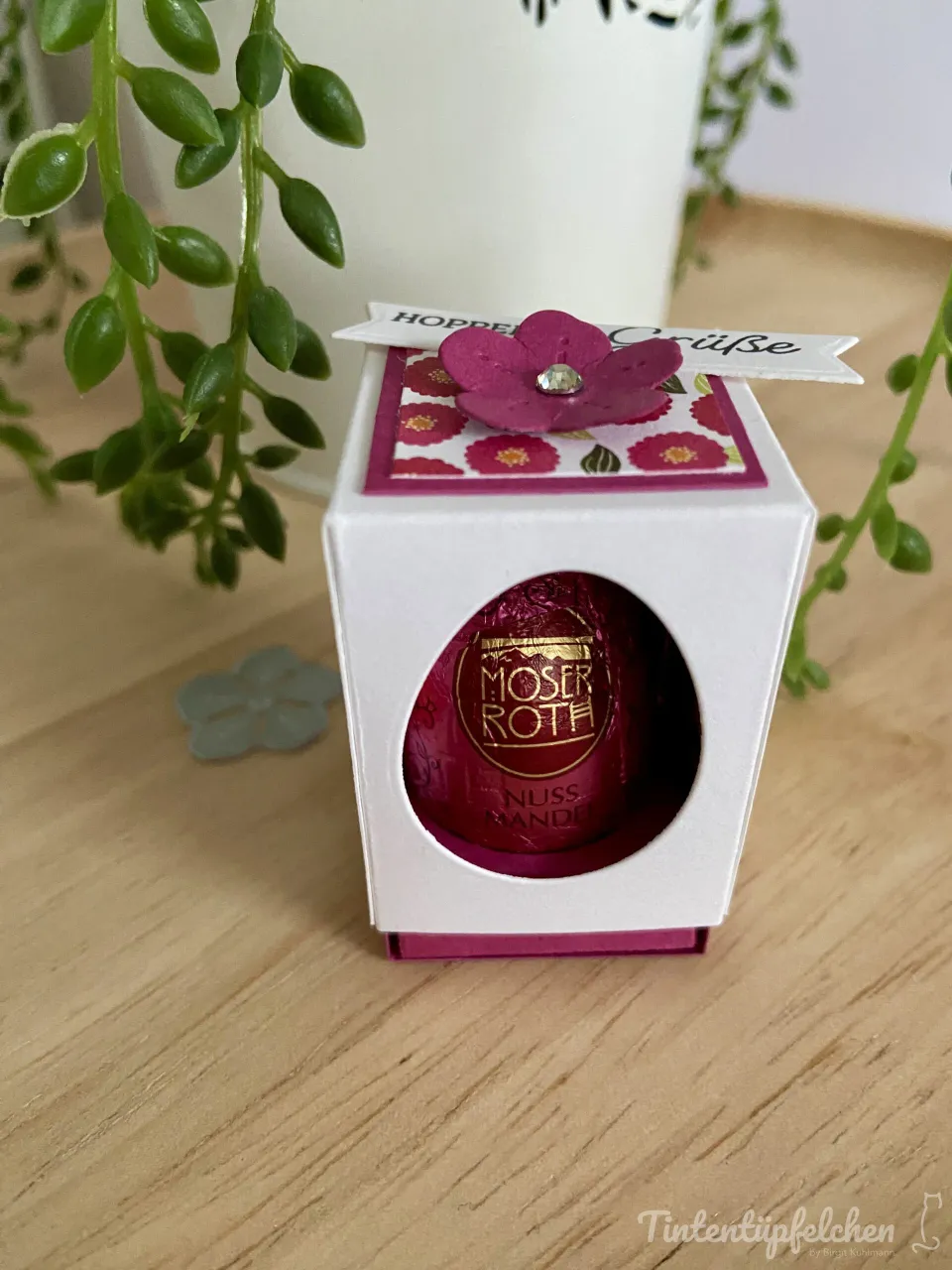 Ein weißer Karton mit ovaler Fensteröffnung zeigt eine Moser Roth Nussmande in rotem Folienpapier. Auf der Decke liegt ein rosa Blüthenstempel mit Diamant, darunter ein Streifen mit „Hoppes Grüße“ und einem floralen Muster aus roten Blüten auf weißem Grund.
