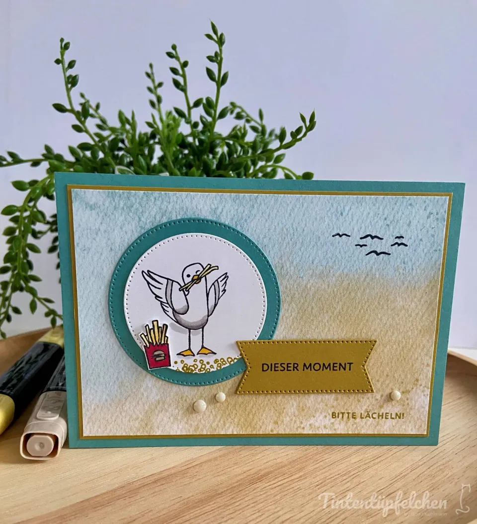 Eine handgebastelte Karte mit aqua-blauem und sandfarbenem Hintergrund, gestempelt mit einem Seeadler, der Nudeln hält. Ein gelber Streifen trägt den Text „Dieser Moment“, unten steht „Bitte lächeln!“. Links sind zwei Marker sichtbar, rechts schweben kleine Silhouetten von Vögeln.