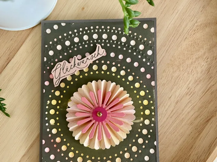 Eine gestempelte Karte mit dunkelgrünem Hintergrund und weißem Punktmuster, zentriert von einer gefalteten Papierblume in Rosa- und Beigetönen mit einem goldenen Mittelpunkt. Oben ist ein Stempel mit der Aufschrift „Glockwunsch“ angebracht.