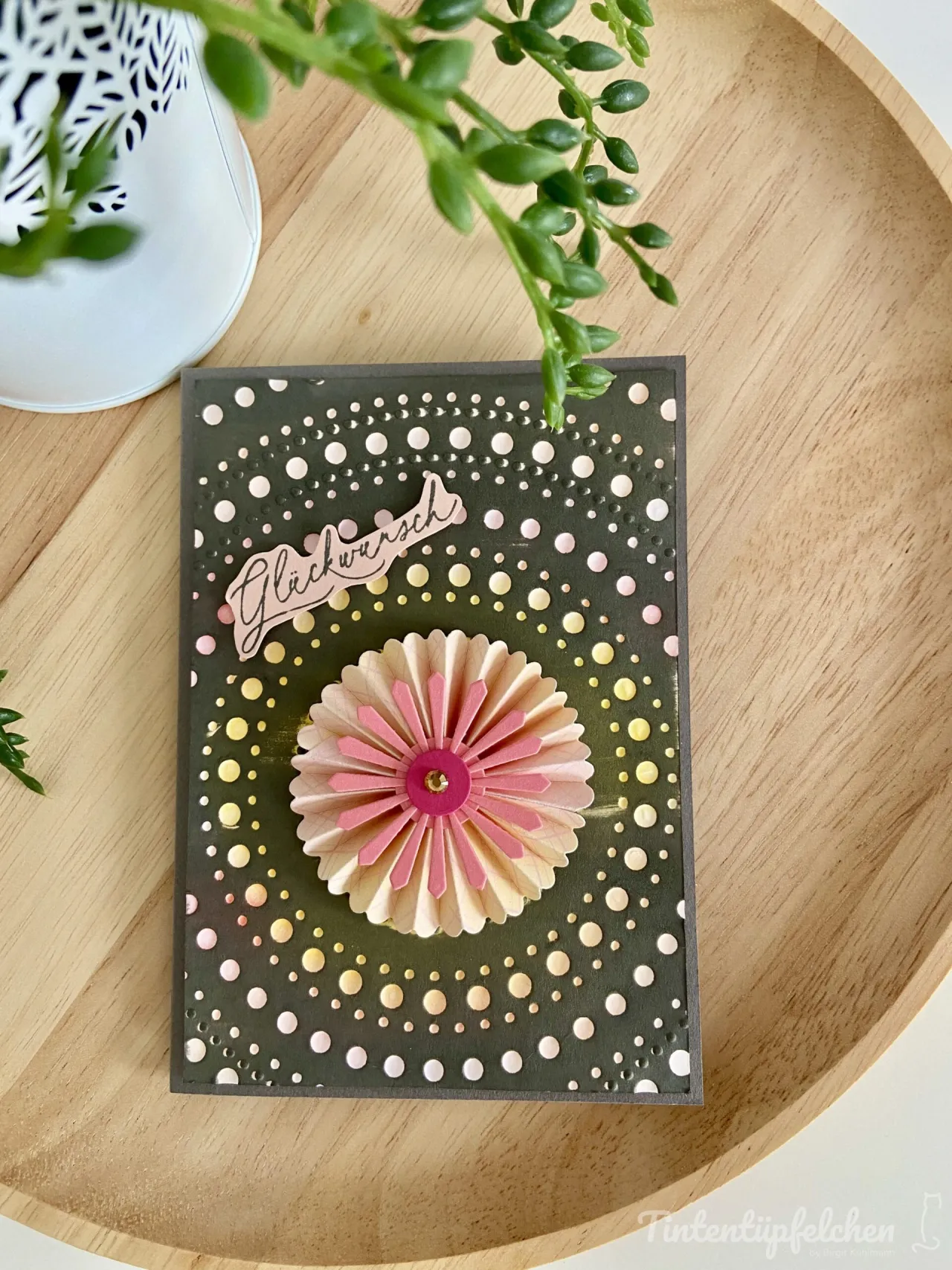 Eine gestempelte Karte mit dunkelgrünem Hintergrund und weißem Punktmuster, zentriert von einer gefalteten Papierblume in Rosa- und Beigetönen mit einem goldenen Mittelpunkt. Oben ist ein Stempel mit der Aufschrift „Glockwunsch“ angebracht.