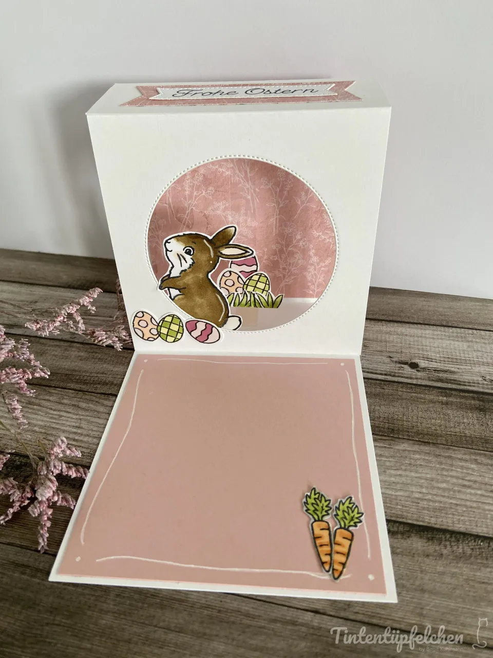 Eine Oster-Karte mit weißem Karton und rosa Hintergrundpapier zeigt einen gestempelten Hasen, der Eier hält, umgeben von weiteren dekorierten Eiern. Zwei Karotten sind unten rechts angebracht. Oben ist ein Band mit der Aufschrift „Frohe Ostern“ sichtbar.
