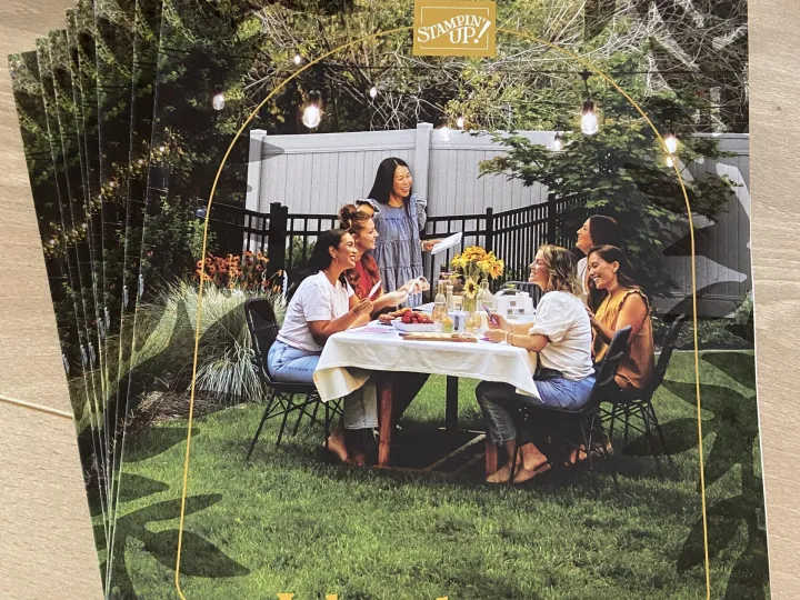 Ein Stapel Jahreskataloge von Stampin' Up! liegt auf einem Holltisch. Das Cover zeigt ein Foto von sechs Frauen, die draußen an einem Tisch mit Sonnenblumen und Kerzen sitzen. Ein weißer Docht serviert den Katalog oben ab.