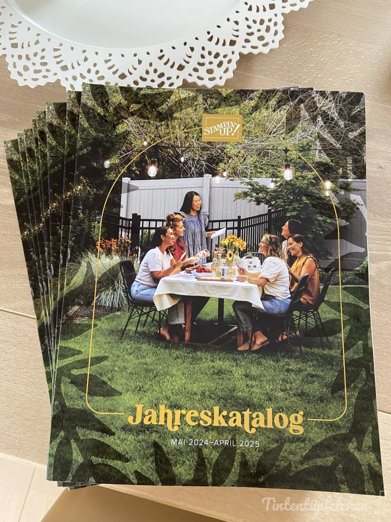 Ein Stapel Jahreskataloge von Stampin' Up! liegt auf einem Holltisch. Das Cover zeigt ein Foto von sechs Frauen, die draußen an einem Tisch mit Sonnenblumen und Kerzen sitzen. Ein weißer Docht serviert den Katalog oben ab.