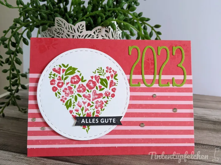 Eine gestempelte Karte mit rotem Grundpapier in weißen Streifen, darauf ein weißer Kreis mit gestickter Naht. Im Kreis ist ein Herz aus roten und grünen Blumenstempeln zu sehen, darunter ein schwarzer Band-Stempel mit der Aufschrift „ALLES GUTE“. Rechts daneben steht die Zahl „2023“ in grünen Metallstanzern, ergänzt durch kleine glänzende Perlen als Verzierung. Im Hintergrund ist eine grüne Zimmerpflanze in einem weißen geschnitzten Topf zu erkennen.