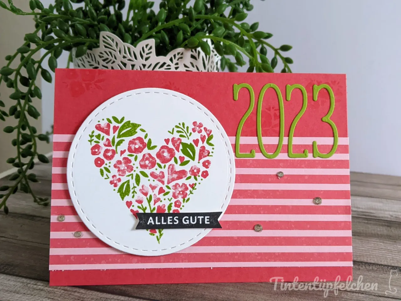 Eine gestempelte Karte mit rotem Grundpapier in weißen Streifen, darauf ein weißer Kreis mit gestickter Naht. Im Kreis ist ein Herz aus roten und grünen Blumenstempeln zu sehen, darunter ein schwarzer Band-Stempel mit der Aufschrift „ALLES GUTE“. Rechts daneben steht die Zahl „2023“ in grünen Metallstanzern, ergänzt durch kleine glänzende Perlen als Verzierung. Im Hintergrund ist eine grüne Zimmerpflanze in einem weißen geschnitzten Topf zu erkennen.