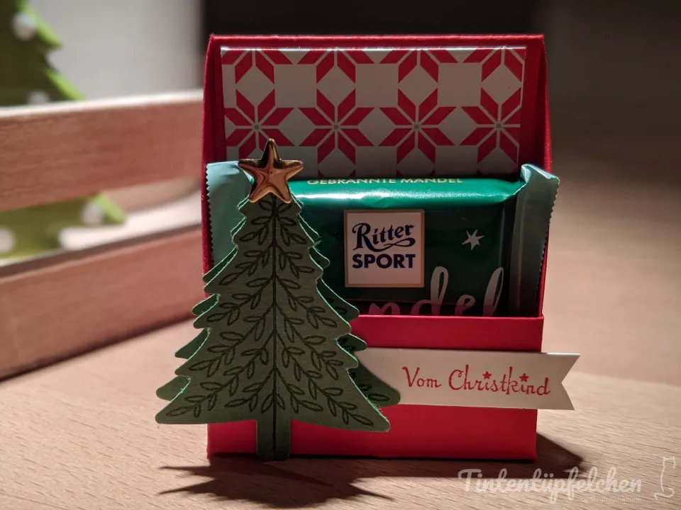 Eine rote Geschenkbox mit einem Ritter Sport Schokoriegel und einem aufgesteckten Weihnachtsbaum aus Papier. Auf der Box ist ein Banner mit der Aufschrift "Vom Christkind" angebracht.