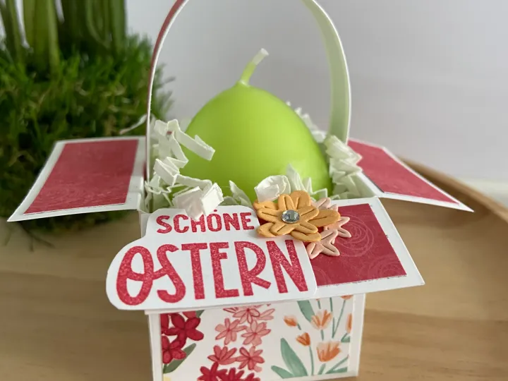 Ein Oster-Kasten aus Papier mit floralen Mustern in Rot, Pink und Grün, der ein grünes Ei enthält. Die Decke zeigt die Beschriftung „Schöne Ostern“ in roten Lettern und ist mit einer gelben Blüte und weißem Papierstreusel verziert.