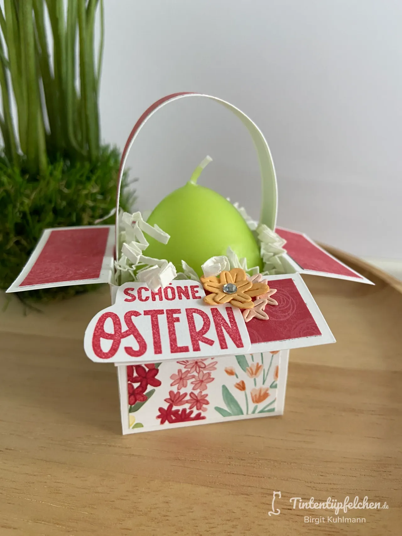 Ein Oster-Kasten aus Papier mit floralen Mustern in Rot, Pink und Grün, der ein grünes Ei enthält. Die Decke zeigt die Beschriftung „Schöne Ostern“ in roten Lettern und ist mit einer gelben Blüte und weißem Papierstreusel verziert.