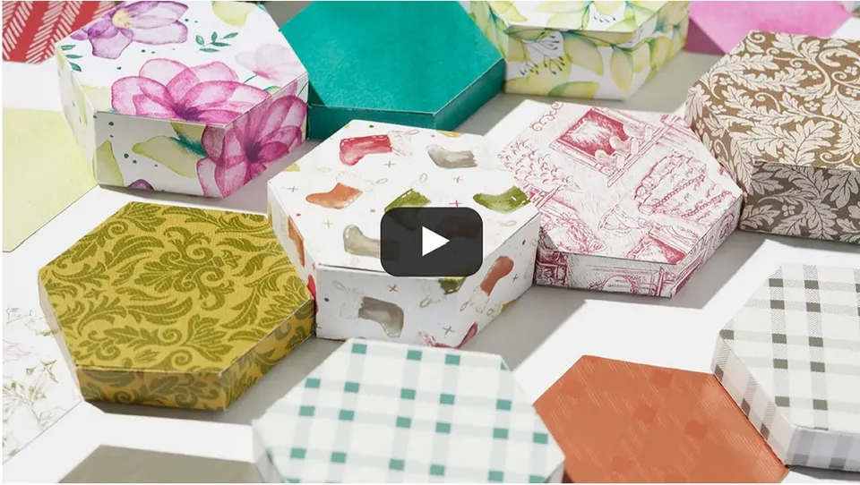 Vielfältige hexagonale Geschenkboxen aus gestempelten und bemalten Papier mit Motiven wie Blumen, Damengarnstilen, Checkern und geometrischen Musten in Farben von Rosa bis Grün. Ein Play-Button deutet auf ein Video hin.