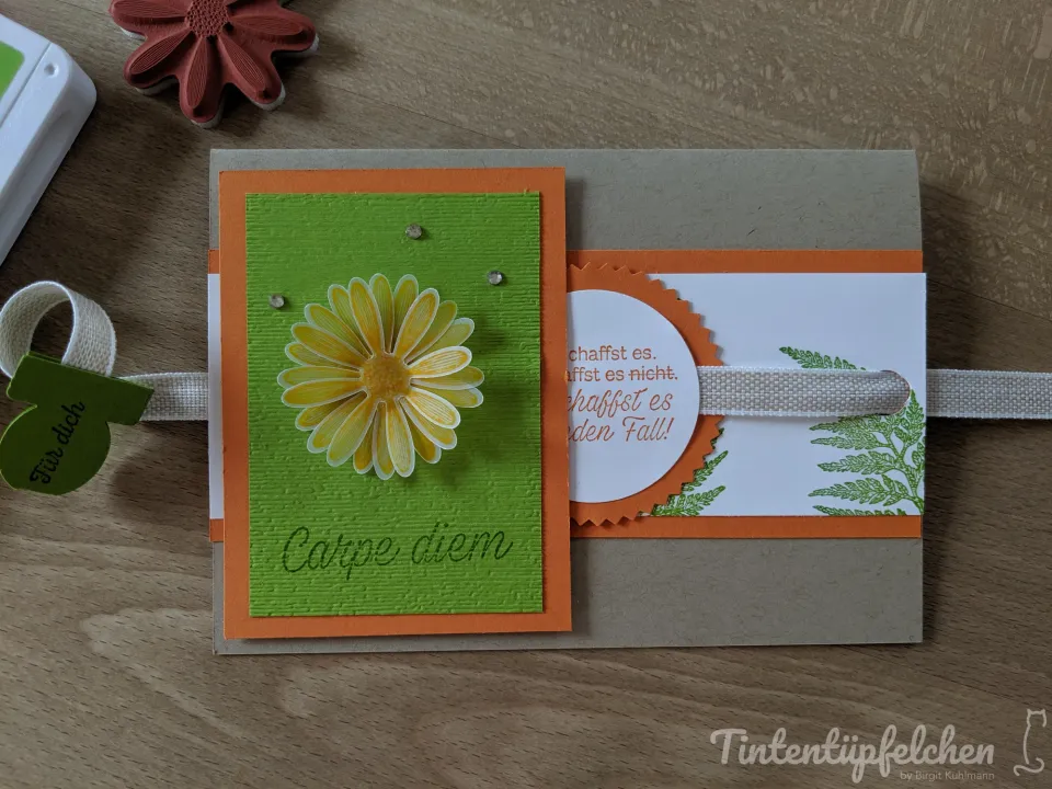 Eine Bastelkarte mit orangefarbenem und grünem Karton, darauf eine gestempelte gelbe Blume und die Aufschrift „Catpe diem“. Ein Bogen mit Textfragmenten ist über einen Stoffstreifen verschiebbar. Im Hintergrund sind ein Stempel und ein grüner Papieranhänger zu sehen.