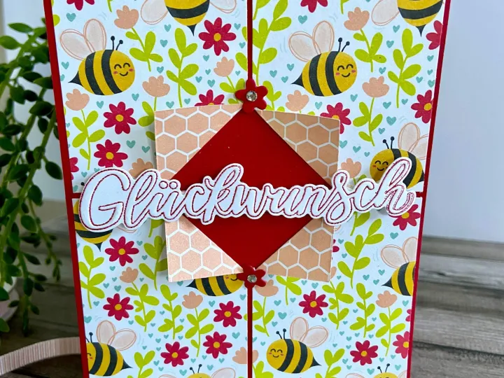 Eine gestempelte Karte mit einem Bienen- und Blumenmuster in Gelb, Rot, Grün und Rosa. Im Zentrum befindet sich ein roter Diamant auf hellrosa Wellenlinienpapier, über dem das Wort „Glückwunsch“ in weißer Schrift mit roten Konturen liegt. Zwei kleine rote Blümchen als Aufnäher schließen die Mitte zusammen.