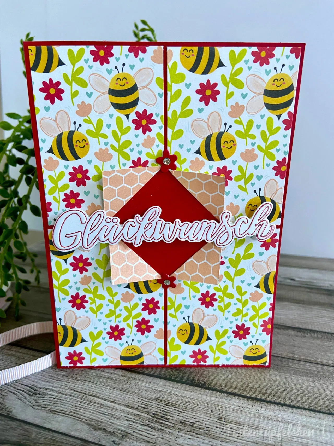 Eine gestempelte Karte mit einem Bienen- und Blumenmuster in Gelb, Rot, Grün und Rosa. Im Zentrum befindet sich ein roter Diamant auf hellrosa Wellenlinienpapier, über dem das Wort „Glückwunsch“ in weißer Schrift mit roten Konturen liegt. Zwei kleine rote Blümchen als Aufnäher schließen die Mitte zusammen.