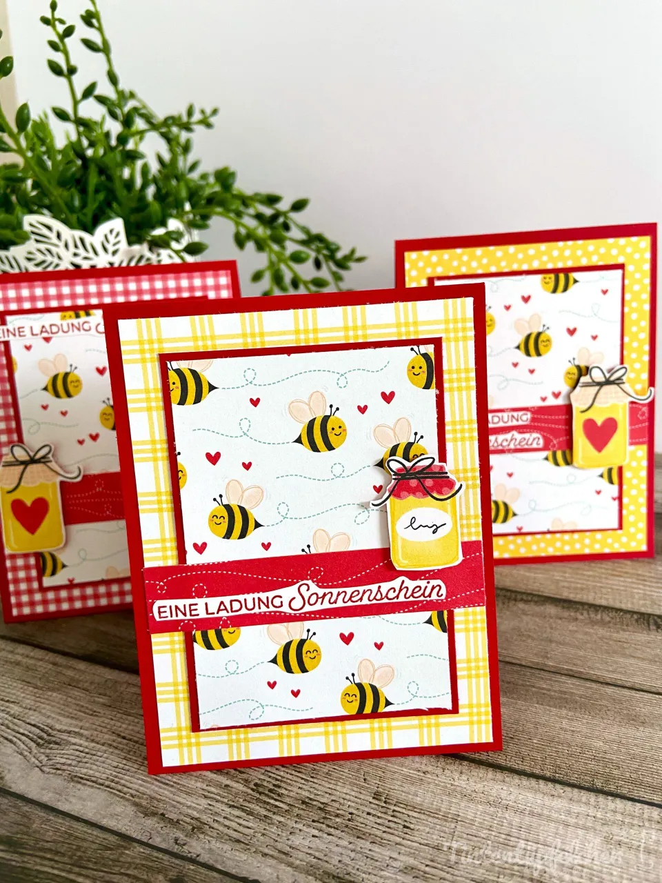 Drei selbstgebastelte Einladungskarten mit Bienen-Motiven in Gelb, Rot und Weiß stehen auf einem Holzuntergrund. Die Karten zeigen gestempelte Bienchen, kleine Herzen und eine 3D-Elemente wie ein Honigglas mit „luz“ Beschriftung. Der Text „EINE LADUNG Sonnenschein“ ist auf roten Streifen gedruckt. Im Hintergrund steht eine grüne Pflanze in einer weißen Schale.