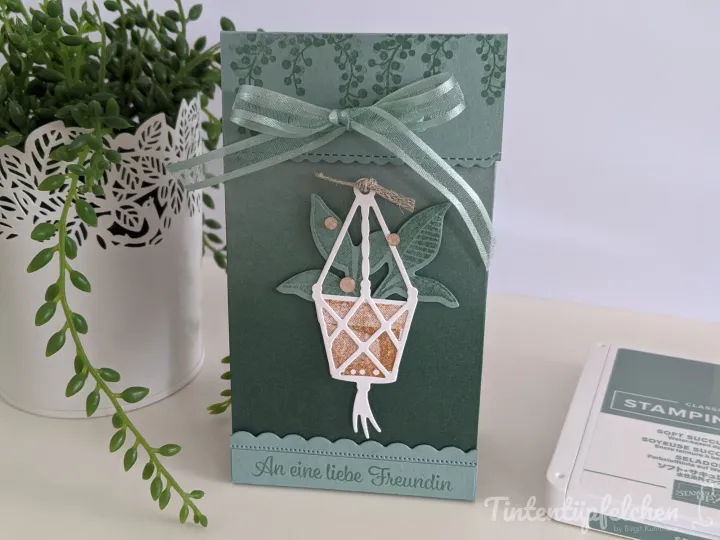 Eine grüne Karte mit gestempeltem Blattmuster und einem weißen Stanzbild eines Hängerkörbsch, gefüllt mit goldfarbenem Glitzerpapier. Ein hellgrüner Seidenband ist oben zu einer Schleife gebunden. Unten steht die Aufschrift „An eine liebe Freundin".