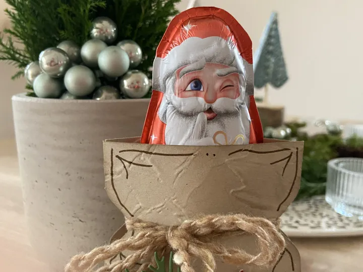 Ein handgefertigter Adventskalender aus braunem Papier mit gestempelten Linien, an dem ein rotes Seil gebunden ist. Davor liegt eine grüne Tannenzweige-Stempelung und ein glitzernder roter Stern mit der Aufschrift „Alles Liebe vom Nikolaus“. Im oberen Teil ragt ein Schokoladenverpackungsbeutel mit einem lachenden Weihnachtsmann-Gesicht hervor. Im Hintergrund steht eine kleine Tanne in einer grauen Vase mit silbernen Kugeln.