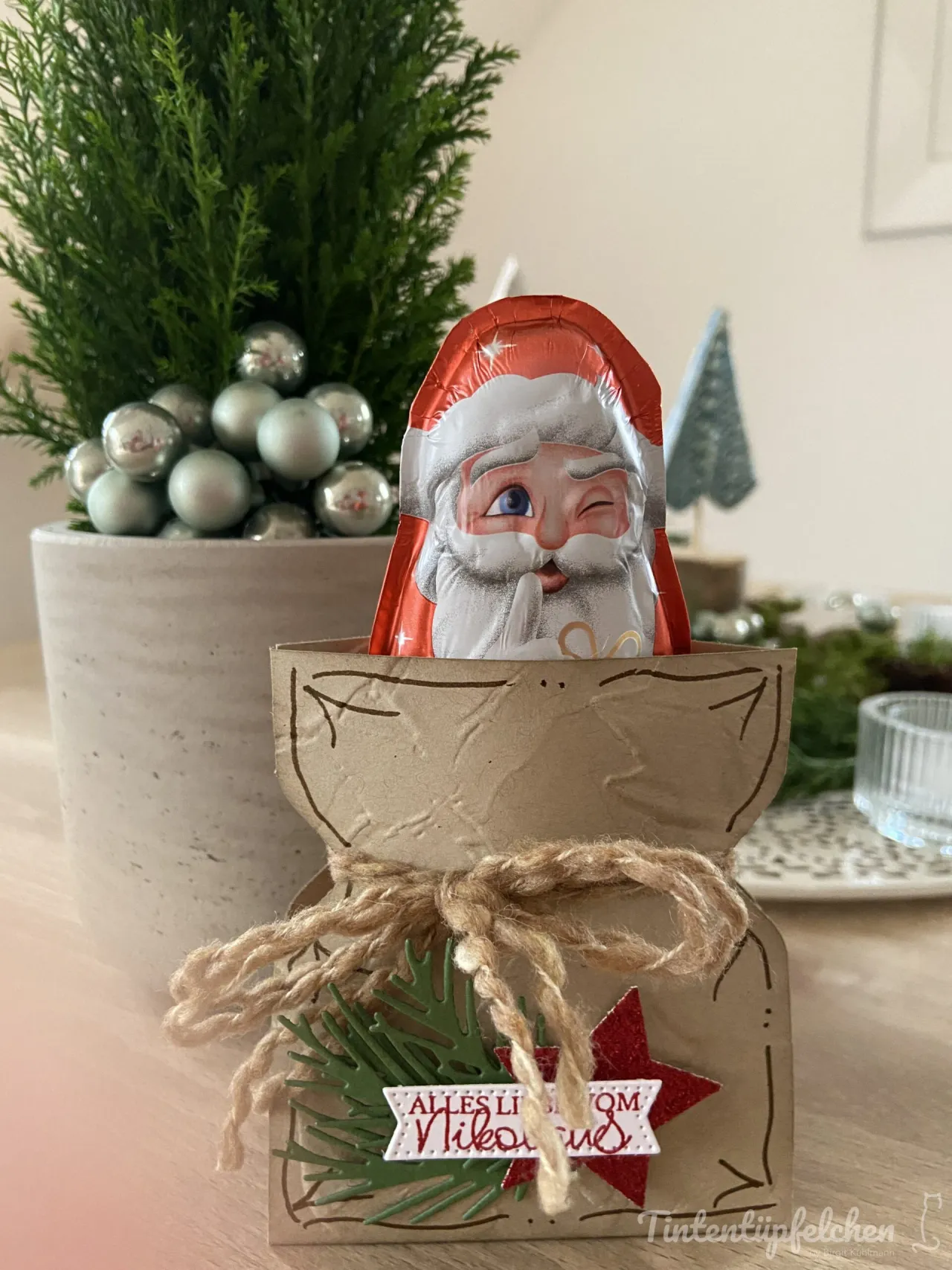 Ein handgefertigter Adventskalender aus braunem Papier mit gestempelten Linien, an dem ein rotes Seil gebunden ist. Davor liegt eine grüne Tannenzweige-Stempelung und ein glitzernder roter Stern mit der Aufschrift „Alles Liebe vom Nikolaus“. Im oberen Teil ragt ein Schokoladenverpackungsbeutel mit einem lachenden Weihnachtsmann-Gesicht hervor. Im Hintergrund steht eine kleine Tanne in einer grauen Vase mit silbernen Kugeln.