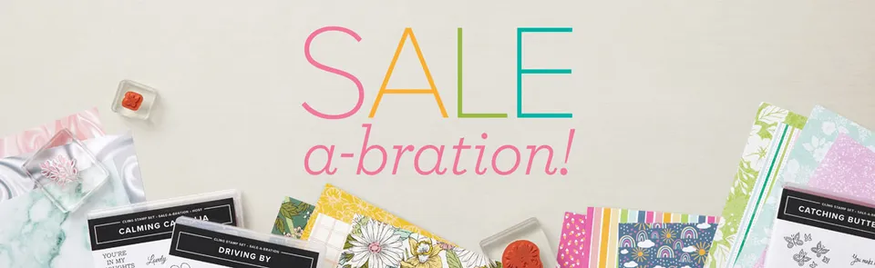 Farbiges Schriftzug „SALE a-bration!" auf weißem Hintergrund, umgeben von Papieren mit Blumen- und Regenbogen-Mustern sowie Stempeln in verschiedenen Farben.