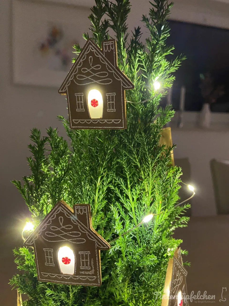 Kleine Haus-Ornamente aus dunkelbraunem Papier mit weißer Linienzeichnung hängen an einer Tannenzweigdeko und leuchten durch integrierte LED-Lichter. Die Fenster sind mit roten Punkten verziert, die Form ähnelt einem Zuckerschmelzhaus.