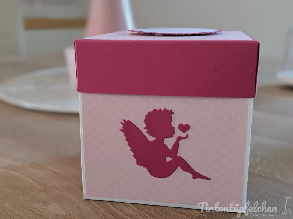 Eine pinke Geschenkbox mit einem Engel-Motiv, das einen Herzluftballon pustet. Der Deckel der Box ist in einem dunkleren Pink gehalten und mit einem Kreis verziert.