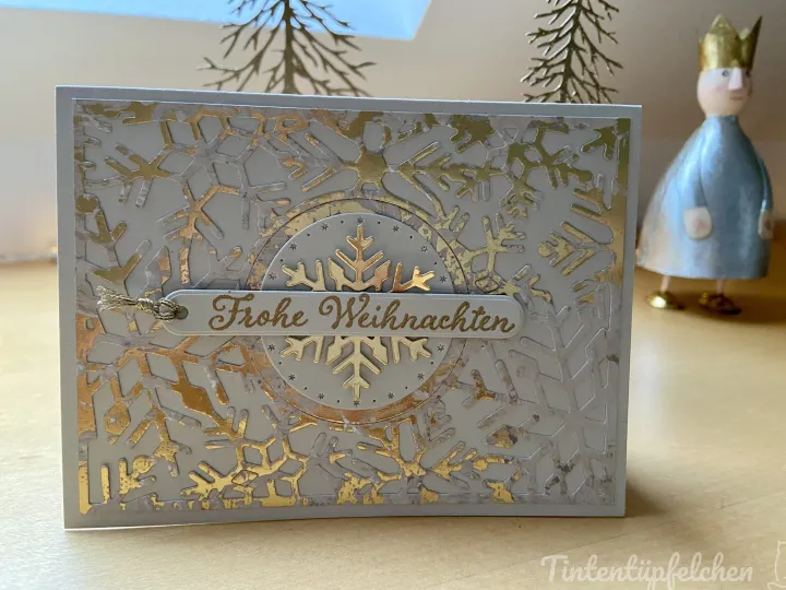 Eine Weihnachtskarte mit silbergrauem Hintergrund und goldenen Schneeflocken-Maschen, die einen zentralen Kreis bilden. Eine goldfarbene Beschriftung „Frohe Weihnachten“ ist auf einem ovalen Element in der Mitte angebracht, umgeben von kleinen Sternen. Im Hintergrund steht ein kleiner Figurenbild mit blauem Gewand und goldenem Kopfschmuck.