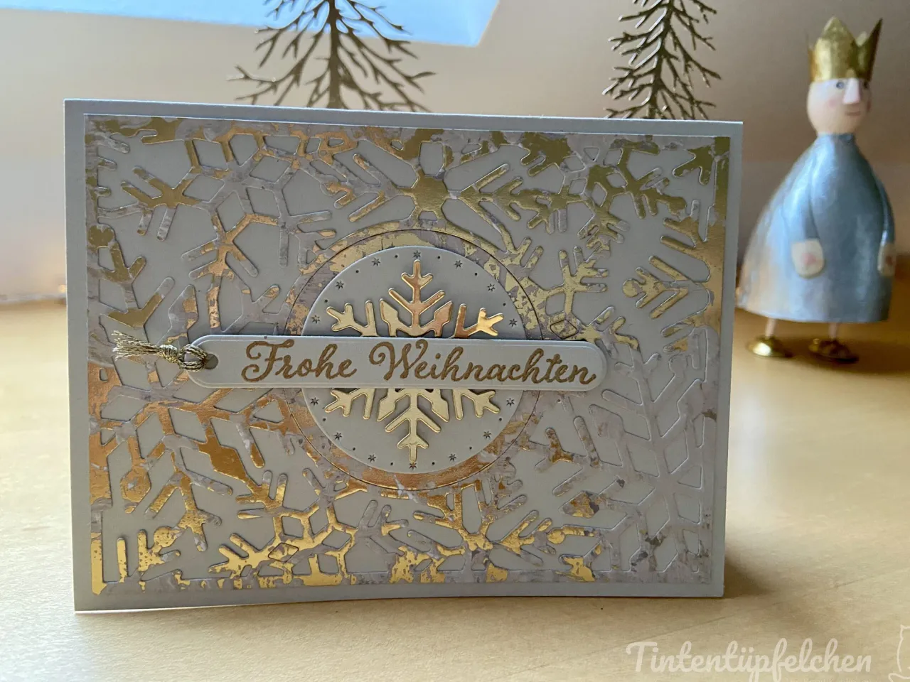 Eine Weihnachtskarte mit silbergrauem Hintergrund und goldenen Schneeflocken-Maschen, die einen zentralen Kreis bilden. Eine goldfarbene Beschriftung „Frohe Weihnachten“ ist auf einem ovalen Element in der Mitte angebracht, umgeben von kleinen Sternen. Im Hintergrund steht ein kleiner Figurenbild mit blauem Gewand und goldenem Kopfschmuck.