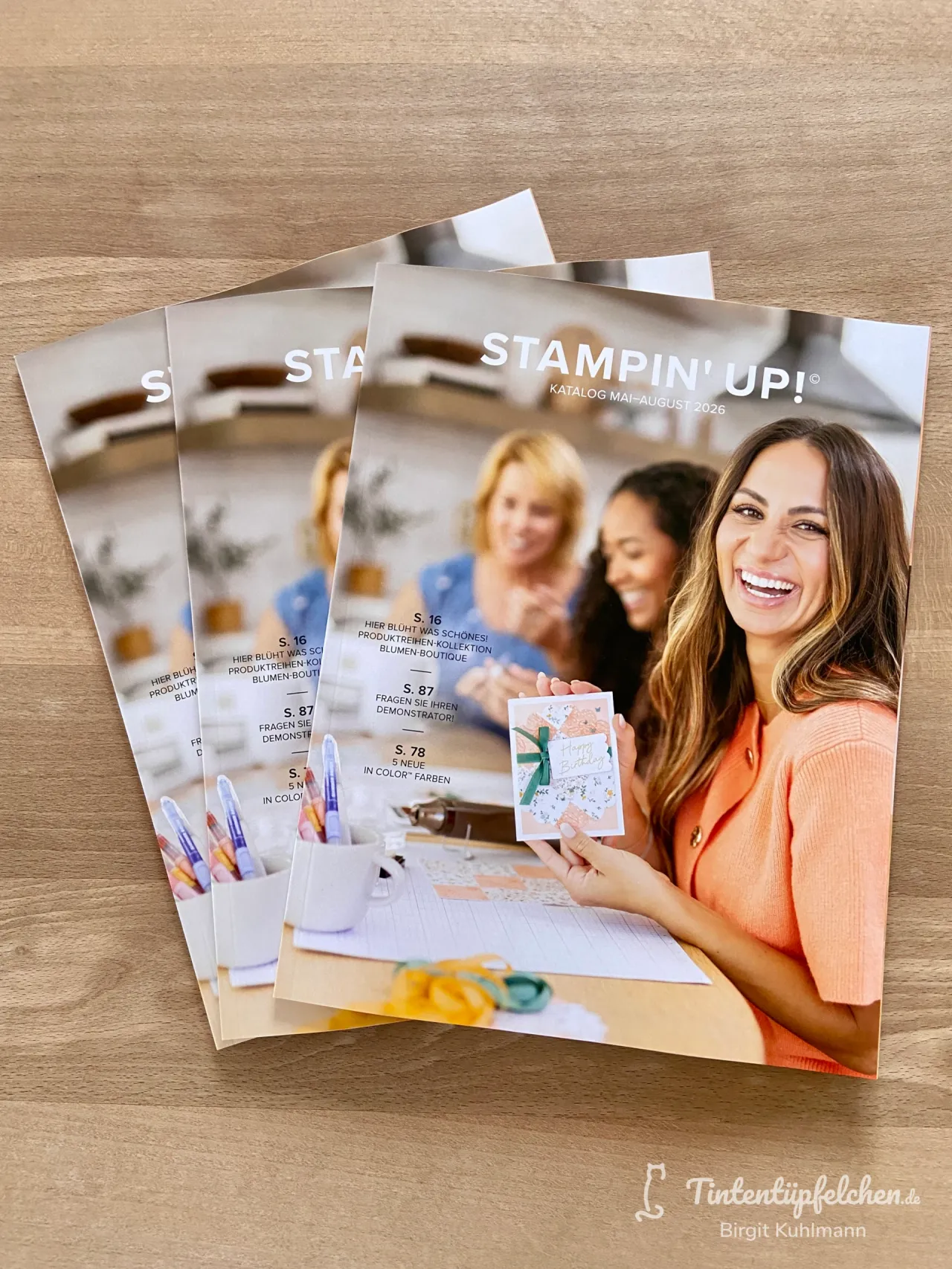 Drei Exemplare des Stampin' Up Katalogs Mai-August 2026 liegen auf einem Holzoberflächen. Der vorderste Katalog zeigt eine lächelnde Frau in orangefarbenem Oberteil, die eine Karte mit grüner Schleife hält; im Hintergrund sind weitere Personen und Bastelmaterial wie Stifte sichtbar.