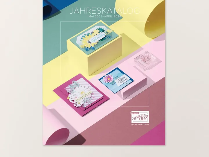 Das Cover des Stampin' Up Jahreskatalogs zeigt gestempelte Karten mit Blumenmotiven in Rosa, Blau und Gelb auf bunten Papierstufen. Der Text „JAHRESKATALOG MAI 2023–APRIL 2024“ ist oben sichtbar, unten steht „AUS DEM JAHRESKATALOG BESTELLEN“. Ein Stempel mit Blumenmuster liegt rechts neben den Karten.