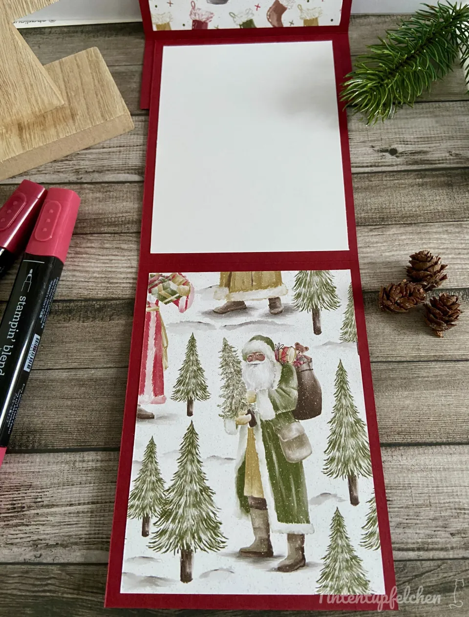 Ein gefalteter Weihnachtsgeschenkkasten mit rotem Rand und einem Stempelbild von Santa Claus in grüner Robe, der Geschenke trägt, umgeben von Tannenbäumen auf schneebedecktem Boden. Links liegen rote Marker und Holzstempel, rechts sind Kiefernzweige und Zapfen als Dekoration platziert.