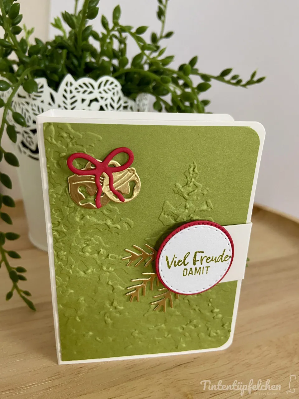 Eine grüne Bastelkarte mit gestempelten Weihnachtsbäumen und Schneeflocken, verziert mit einem roten Papierband als Schleife und zwei goldenen Glocken. Ein weißer Kreis mit rotem Rand zeigt den Text „Viel Freude DAMIT“ in goldener Schrift.