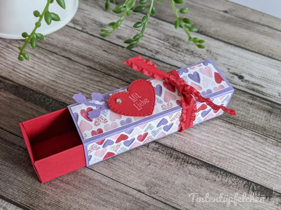 Ein gebasteltes Schachtel-Format aus Papier mit Herz-Muster in Rot, Pink und Lila liegt auf Holzoptik; eine rote Schleife ist um den Deckel gebunden, ein roter Herzmotiv mit der Aufschrift „Mit Liebe" ist als Aufkleber angebracht. Im Hintergrund sind grüne Pflanzenteile sichtbar.