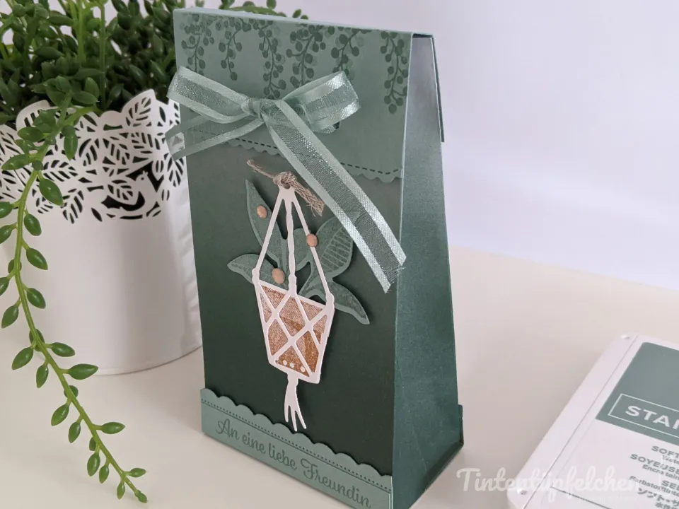 Ein grüner Papiertaschentopf mit gestempeltem Blattmuster und einem gebundenen Seidenband, an dem ein aus Papier ausgeschnittenes Hängeschlösser-Motiv mit Perlen befestigt ist. Unten steht der Text „An eine liebe Freundin" in einer serifenlosen Schrift auf zerknittertem Papier.