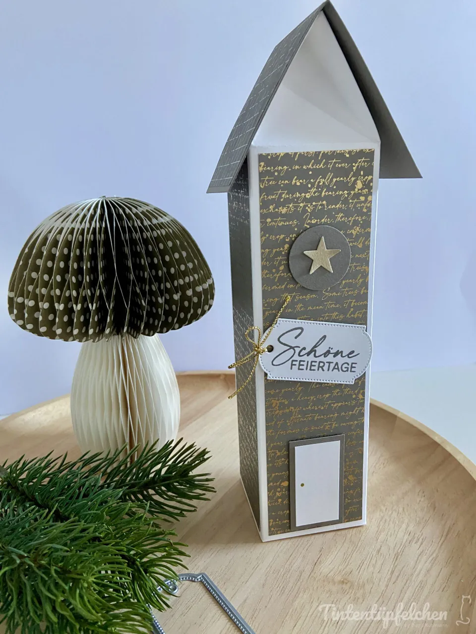 Ein aus Papieren gefertigtes Haus mit grauem Dach und goldschimmerndem Schriftmuster steht auf einem Holzbrett neben einer gefalteten Papierpilzfigur in Grün-Weiß-Muster sowie frischen Tannenzweigen. Ein goldenes Band ist an eine weiße Beschriftungsbuchstabe gebunden, die „Schöne Feiertage“ sagt.