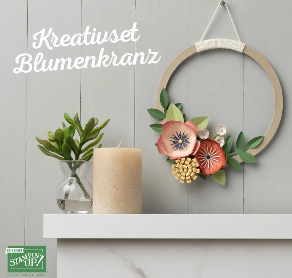 Ein handgebastelter Blumenkranz aus Papier in warmen Tönen wie Rosa, Orange und Gelb hängt an einem Holzring mit geflochtenem Seidenband. Dazu gehören grüne Blattmotive und kleine Stempelblumen auf weißem Hintergrund. Links daneben steht eine Kerze in Beige neben einer kleinen Sukkulentenpflanze in Glasvase vor grauer Wand.