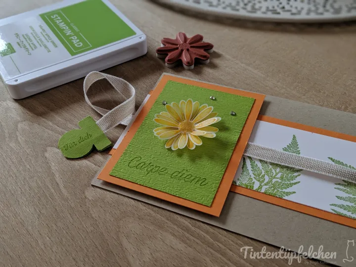 Eine Karte mit grüner Textur und gelbem Daisymotiv liegt auf einem braunen Umschlag, umgeben von einem Stampin' Pad, einer Stempelstift und kleinen Dekorationselementen wie einem Blatt-Schnitt. Die Schrift „Carpe diem“ ist in der Mitte gestempelt, daneben ein weißer Streifen mit grünem Moosmuster.