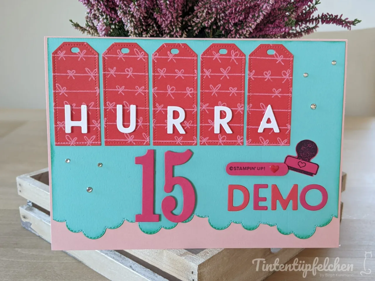 Eine gestanzte Karte mit fünf roten Etiketten, die den Text „HURRA 15 DEMO“ in weißer und roter Schrift tragen. Der Hintergrund ist türkisblau mit einem rosa Wellenrand unten, dekoriert mit kleinen silbernen Glitterpunkten und einem Stempelabdruck von Stampin’ Up! Oben zeigt eine Blumengruppe aus heidriger Purpurfarbe.