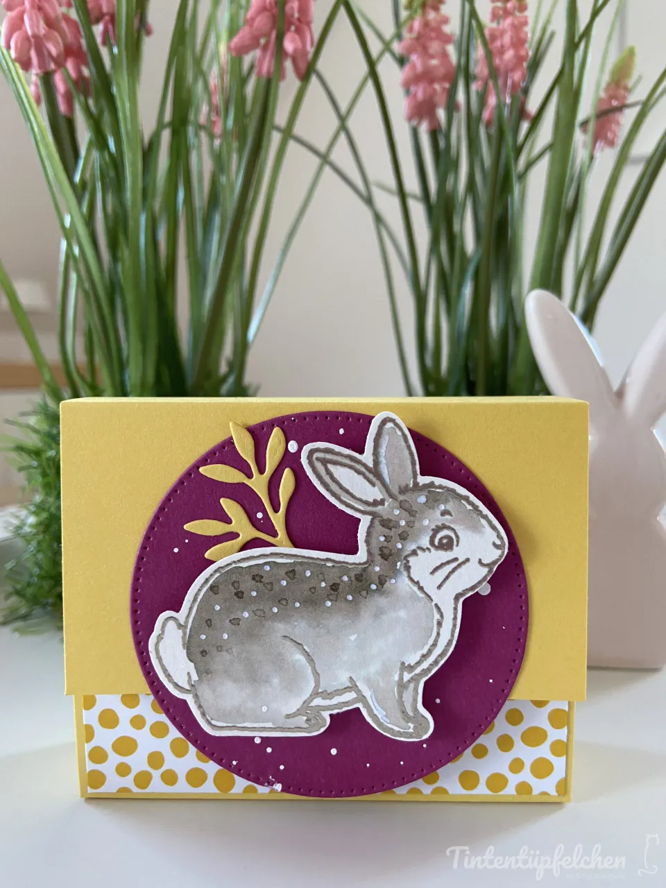 Eine gestempelte Karte mit gelbem Karton, auf dem ein purpurner Kreis mit einem grau getönten Hasenmotiv angebracht ist. Der Hase zeigt kleine weiße Punkte und eine goldfarbene Blütendekoration im Hintergrund des Kreises. Unten befindet sich ein Streifen mit gelben Punkten auf weißem Grund. Im Hintergrund stehen grüne Pflanzenstiele mit rosa Blütenblättern, rechts ist ein Teil eines weißen Hasenfigurins sichtbar.