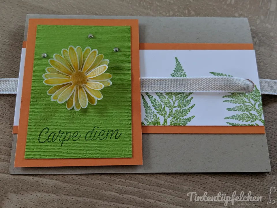 Eine geschmückte Karte mit orangefarbenem Rand und grünem gestempelten Blatt, darauf eine gelbe Blume mit kleinen Glitzersteinen. Der Text „Carpe diem“ ist in schwarzer Schrift auf dem grünen Papier zu sehen. Eine weiße Leinwand-Band läuft quer über die Karte.