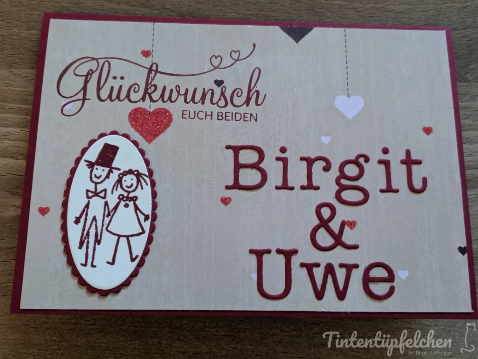 Ein selbstgebasteltes Glückwunschkärtchen in Beige mit dunkelrotem Rand, gestaltet mit gestempelten Texten „Glückwunsch ECH BEIDEN“ und „Birgit & Uwe“. Ein ovaler Ausschnitt zeigt eine stilisierte Zeichnung eines Paares in Hochzeitskleidung. Kleine Herzen aus Glanzpapier hängen als Dekoration an unsichtbaren Fäden, weitere sind verstreut auf dem Hintergrund angebracht.