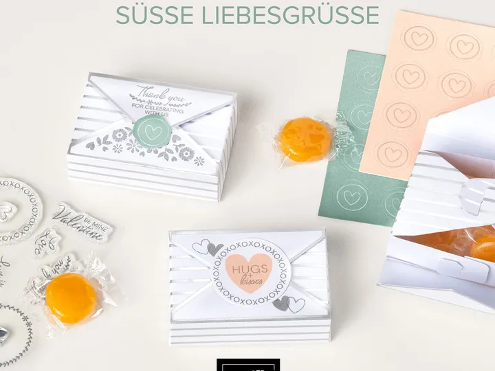 Ein Bastelset mit weißen Umschlag-Karten und gestreiften Boxen, verziert mit grünen und orangefarbenen Stempeln sowie gelben Süßigkeiten in Folie. Die Karte zeigt einen zentralen Kreis mit „HUGS + kisses“ und umgebenden X-Motiven, daneben sind weitere Stempelblätter mit Herzen und Texten wie „Thank you“ sichtbar.
