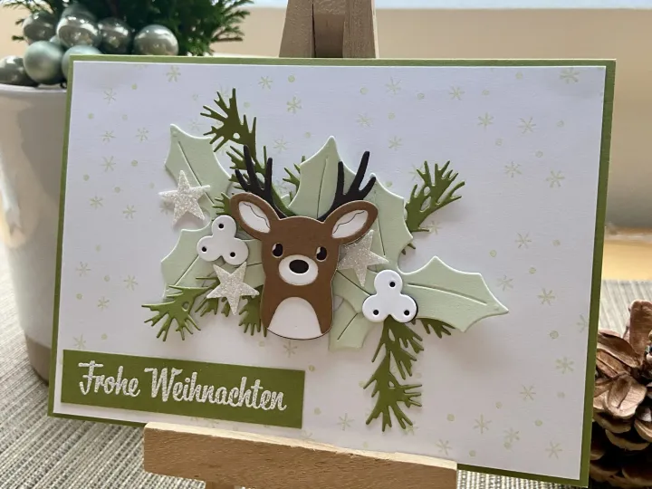 Eine Weihnachtskarte mit einem gestempelten Rehgesicht in Braun und Weiß, umgeben von grünen Eichenblättern und Tannenzweigen. Silberne Sterne und weiße Schleifen als Dekorationen sind eingebettet. Der Text „Frohe Weihnachten“ steht in weißer Schrift auf grünem Papier im unteren Bereich.