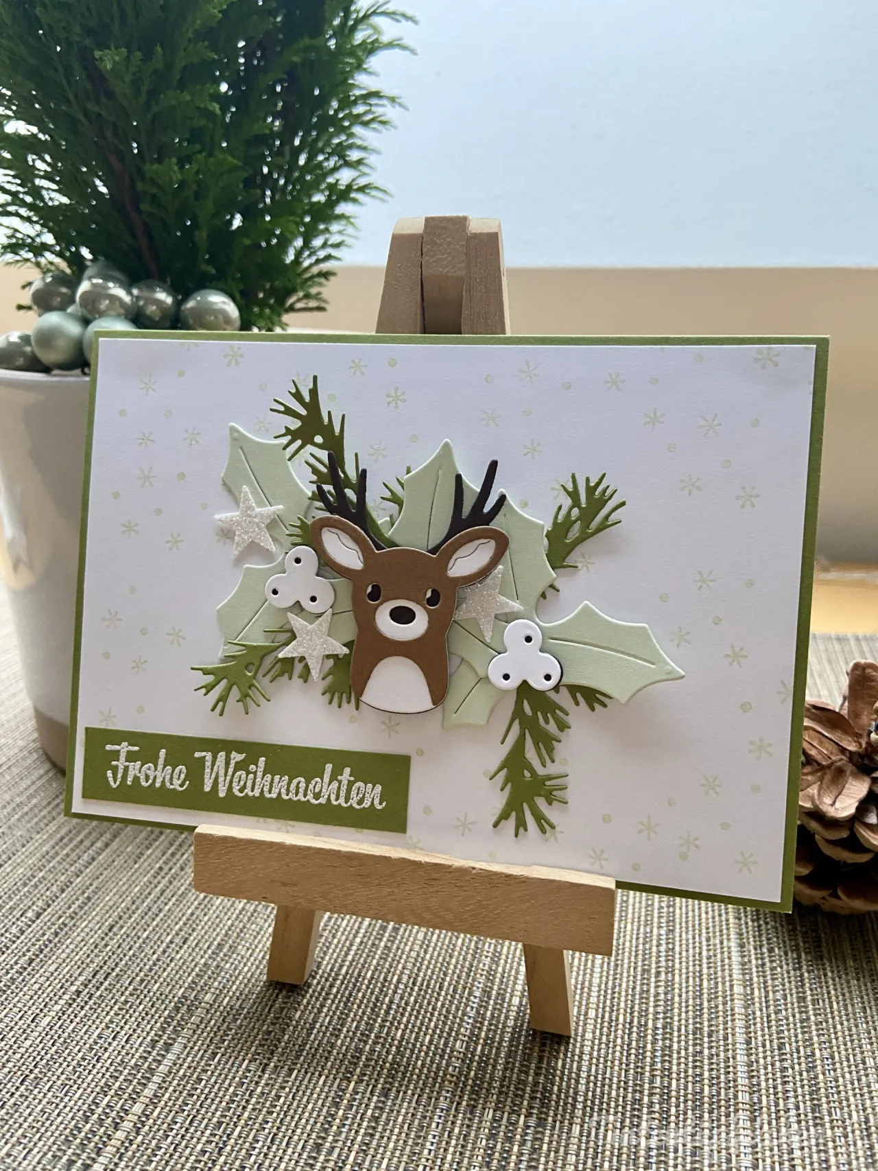 Eine Weihnachtskarte mit einem gestempelten Rehgesicht in Braun und Weiß, umgeben von grünen Eichenblättern und Tannenzweigen. Silberne Sterne und weiße Schleifen als Dekorationen sind eingebettet. Der Text „Frohe Weihnachten“ steht in weißer Schrift auf grünem Papier im unteren Bereich.